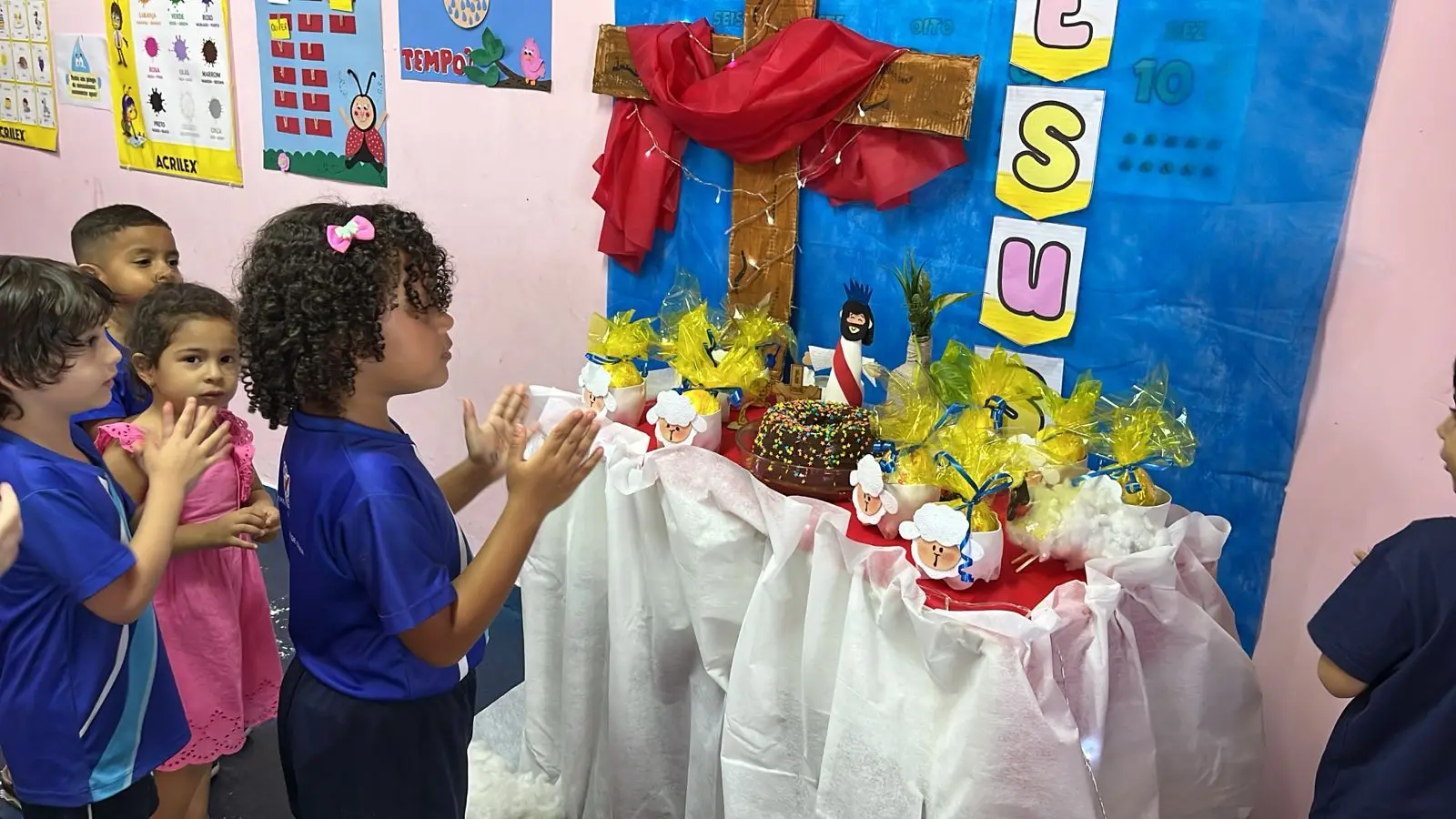 Crianças em pé diante de uma mesa com toalha branca, cruz com pano vermelho e flores amarelas, na sala de aula