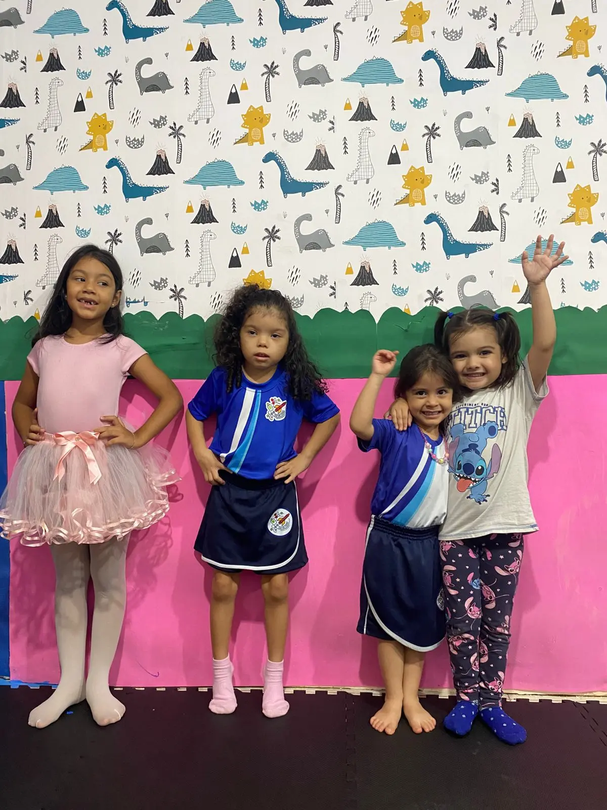 Quatro meninas diante da parede de dinossauros e do painel rosa: uma de collant e saia de tule rosa, as outras de uniforme ou roupa do dia a dia, uma com os braços para o alto
