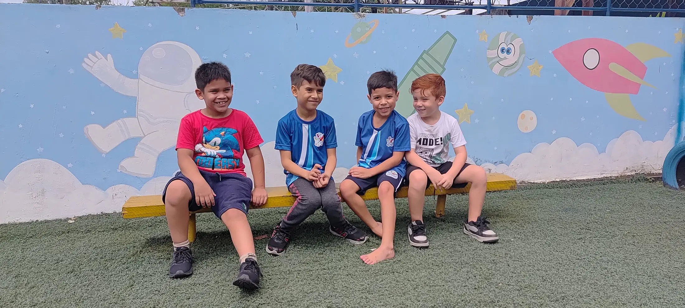 Quatro meninos sentados num banco amarelo do pátio, diante do mural com astronauta e foguete