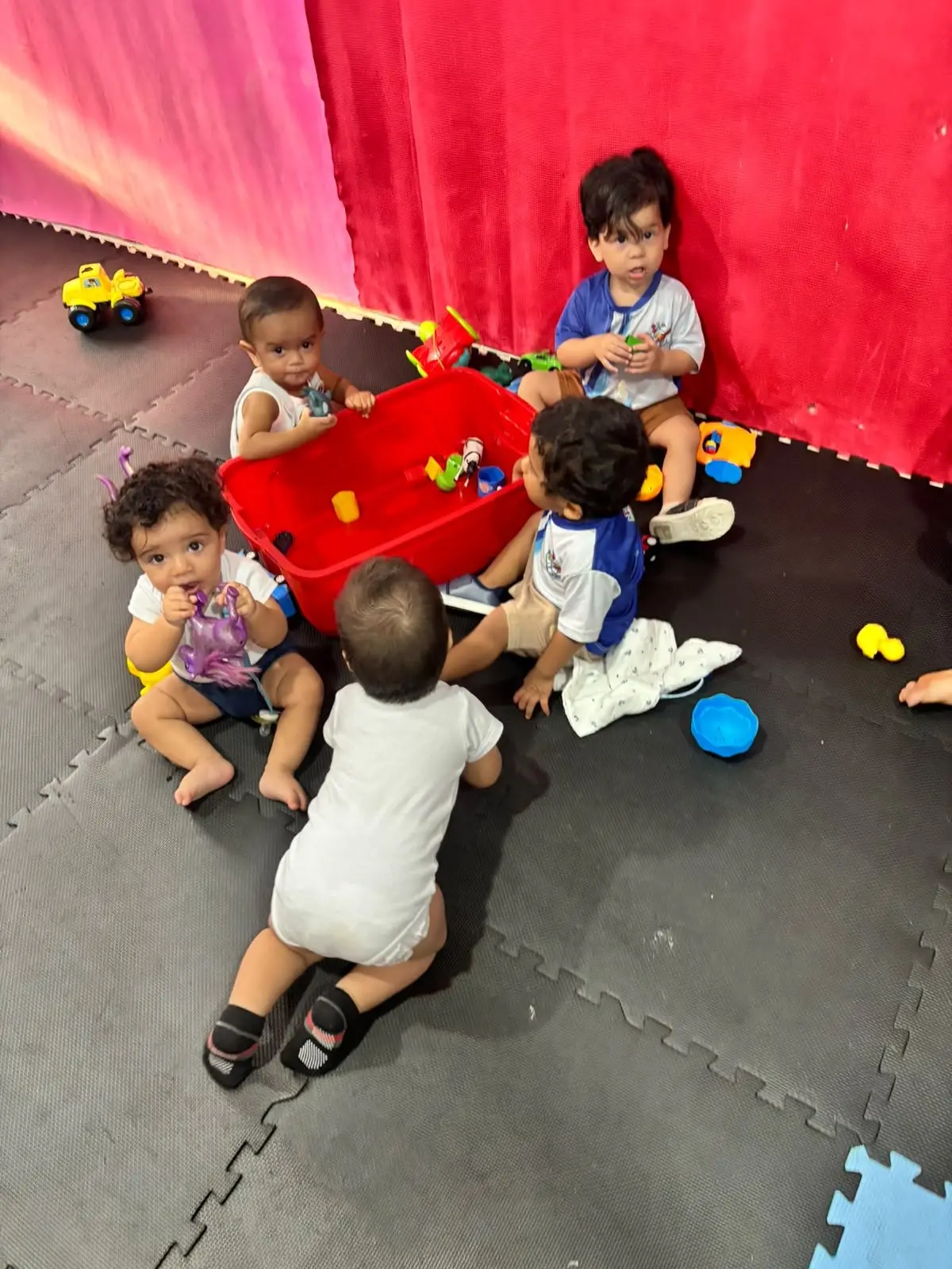 Bebês engatinhando e brincando em volta de uma caixa vermelha cheia de brinquedos