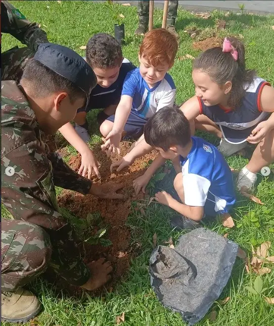 Crianças plantando uma muda no gramado, com um militar fardado ajudando a cavar
