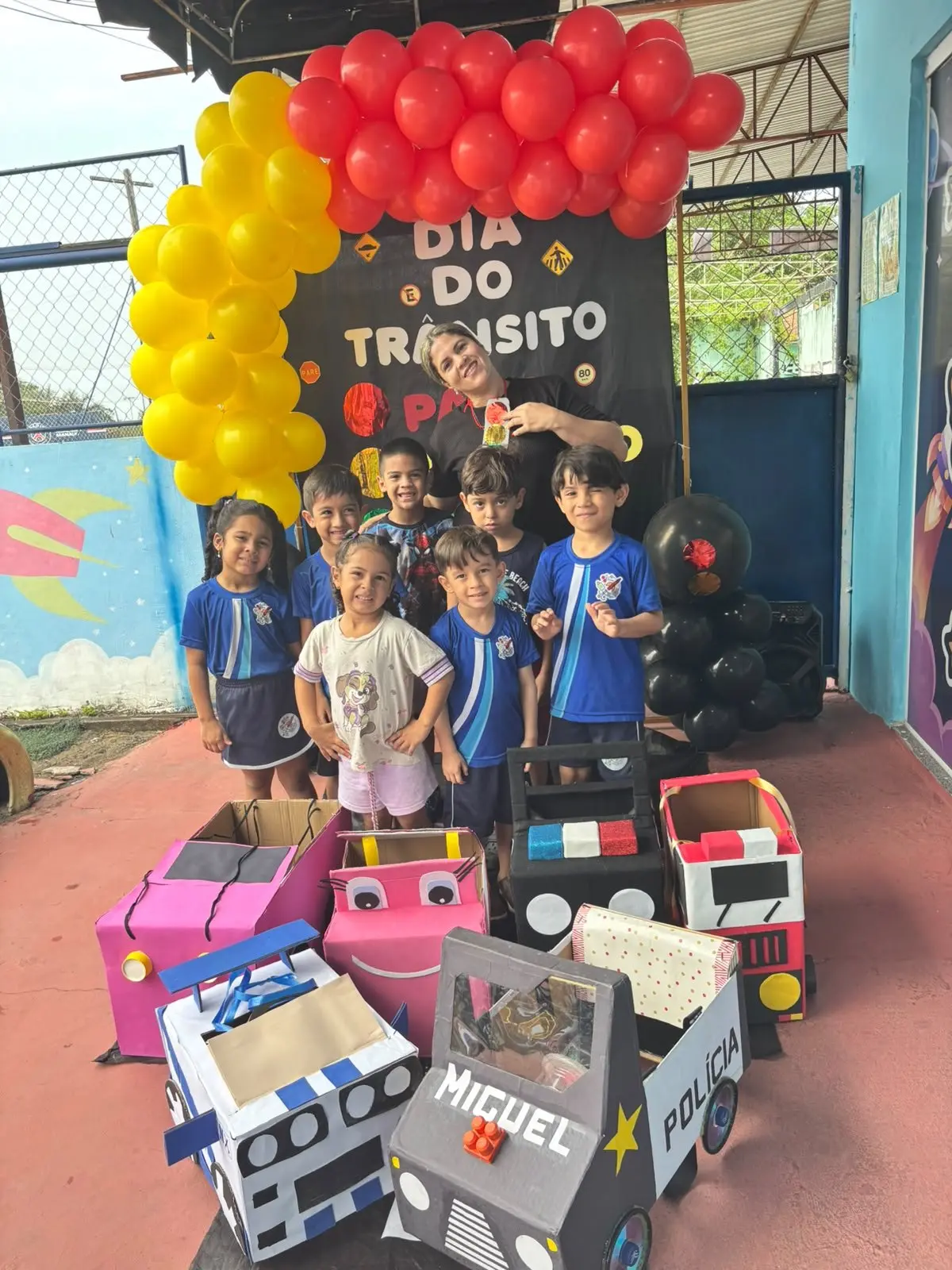 Turma na frente do arco de balões amarelos e vermelhos do Dia do Trânsito, com carrinhos de papelão no chão