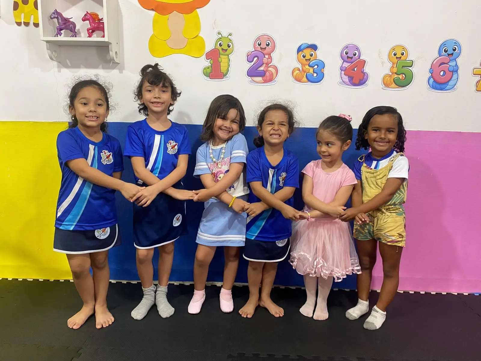 Seis crianças da Educação Infantil de mãos dadas, na frente do mural de números