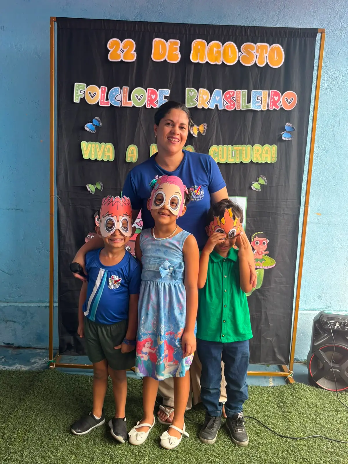 Três crianças de máscara e uma professora de uniforme na frente do painel 22 de agosto, Folclore Brasileiro