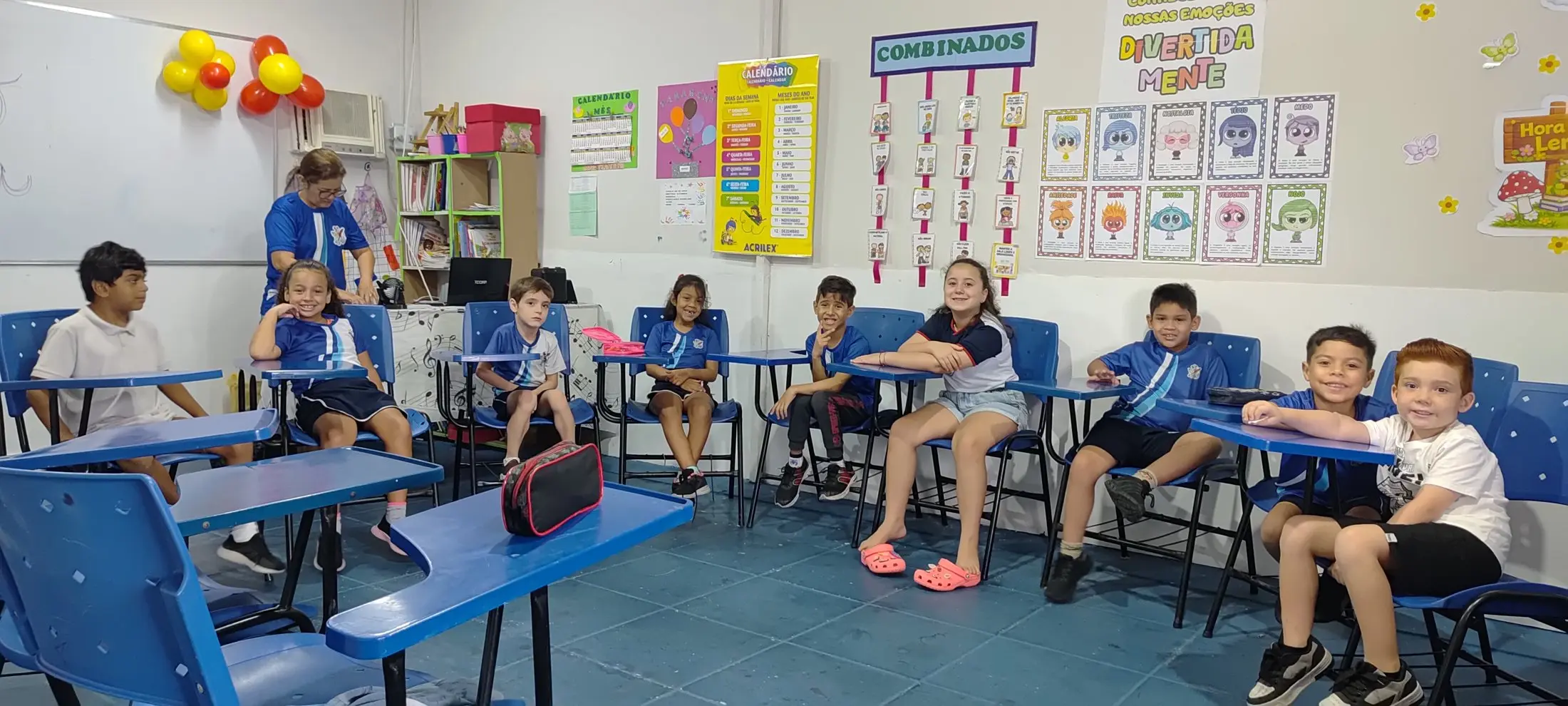Sala de aula com a turma sentada nas carteiras azuis e cartazes coloridos na parede