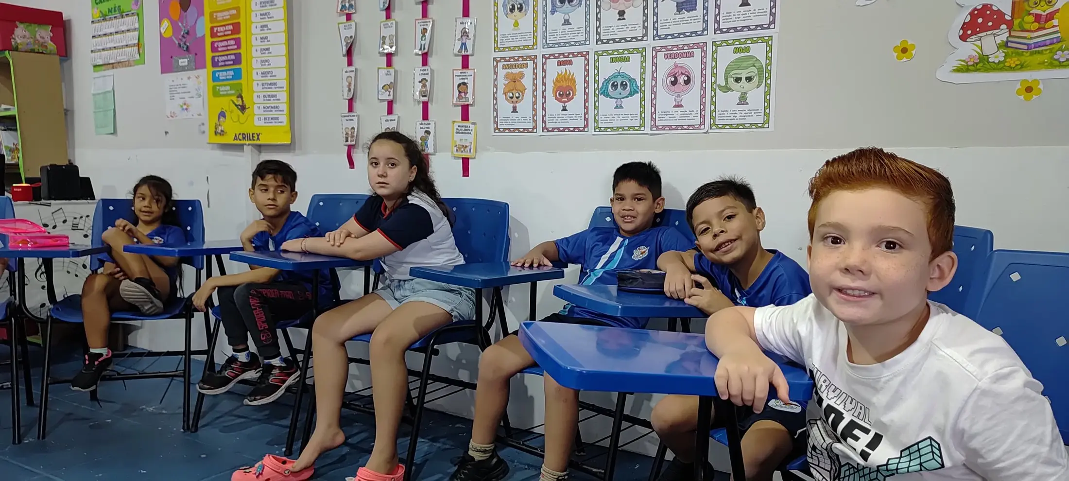 Alunos do Ensino Fundamental sentados nas carteiras azuis, um menino sorrindo em primeiro plano