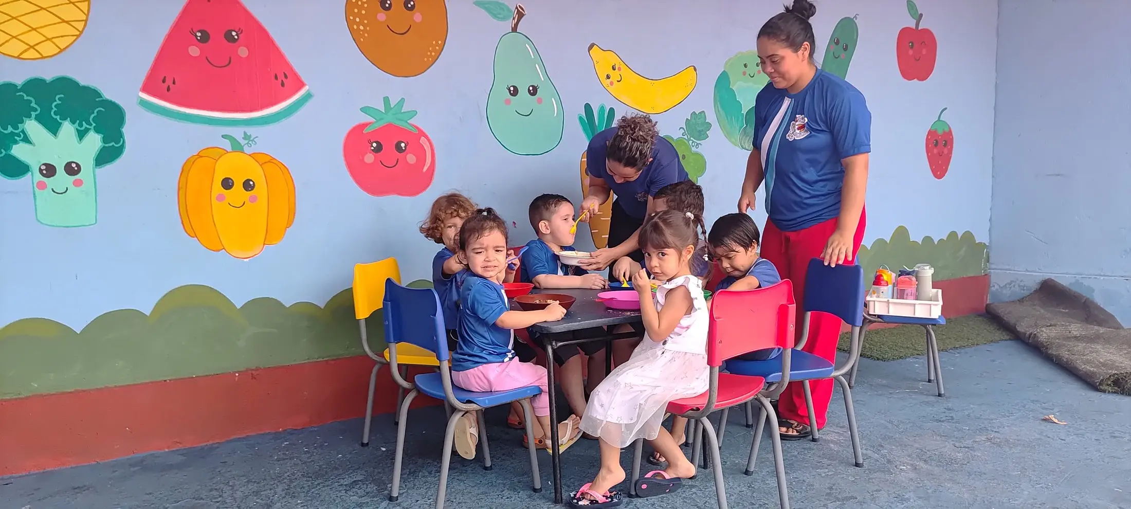 Crianças do Maternal à mesa com a professora, diante do mural de frutas