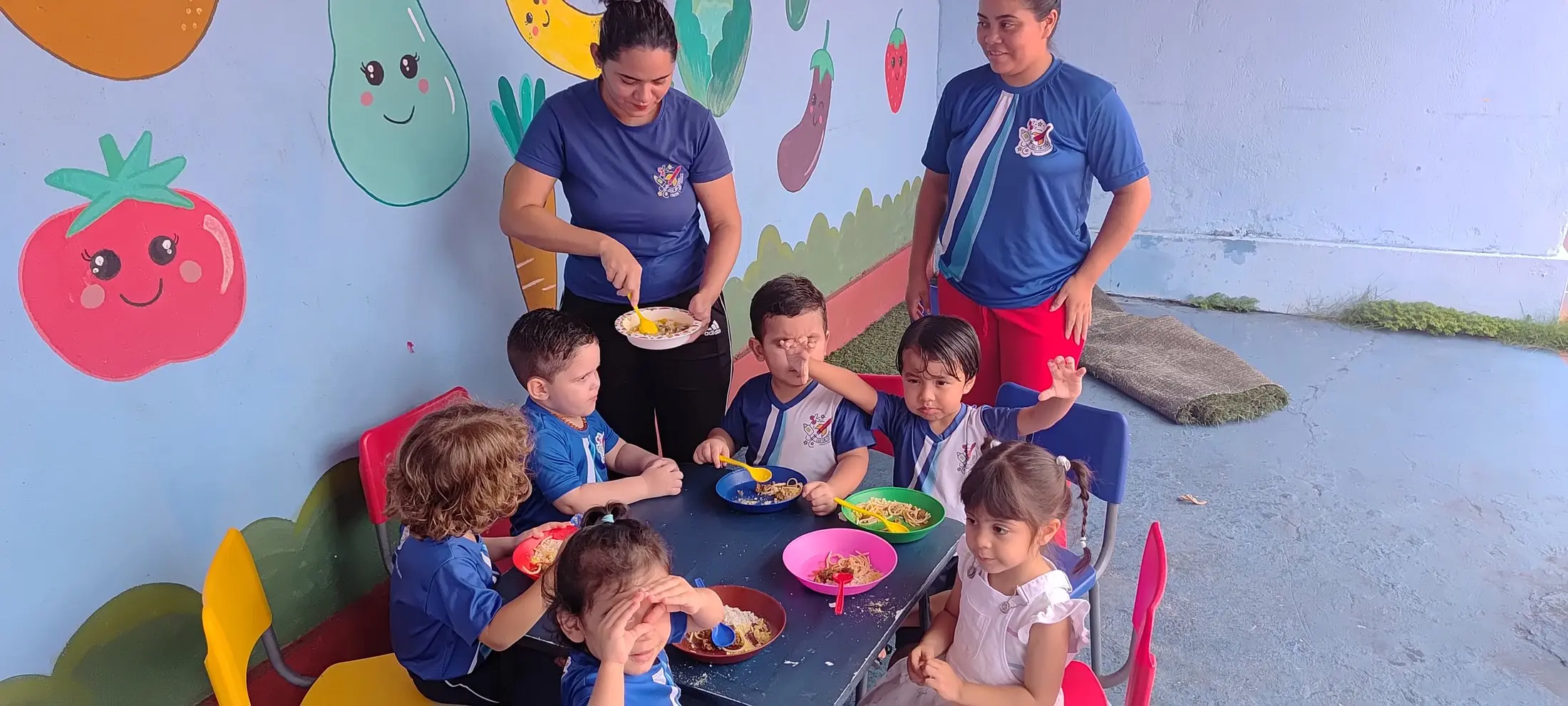 Professora servindo a refeição à turma, com outra ao lado, diante do mural de frutas