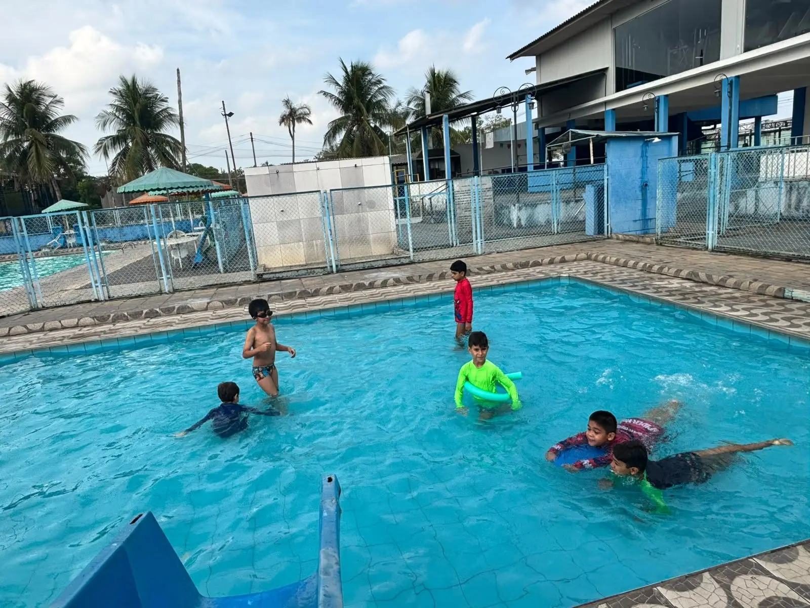 Crianças brincando na piscina com boias coloridas, com coqueiros e o céu ao fundo