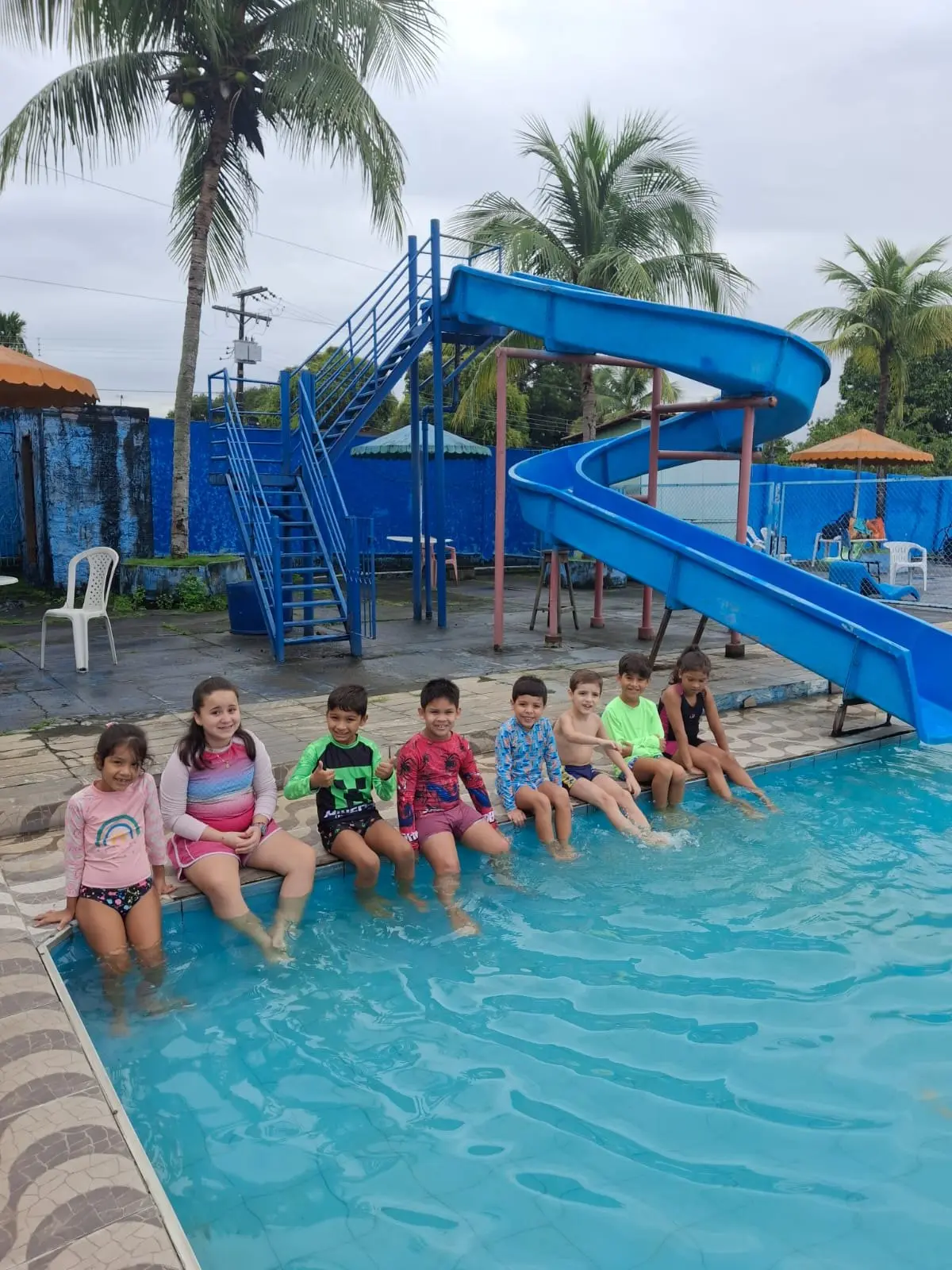 Crianças sentadas na borda da piscina, ao lado do escorregador azul